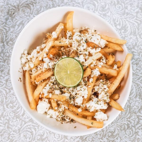 Golden french fries topped with crumbled feta cheese, dried herbs, and a lime slice in a white bowl.