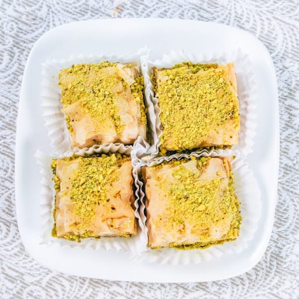 Four square baklava pastries with crushed pistachios on a white plate.