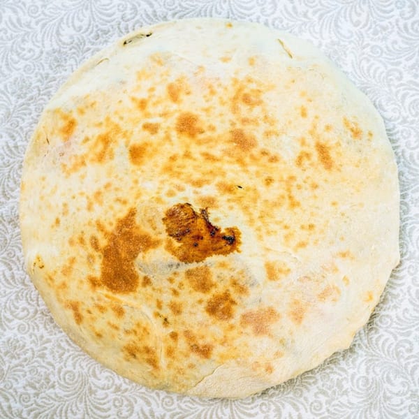 A round, golden-brown flatbread with a slightly charred spot, resting on a patterned white tablecloth.