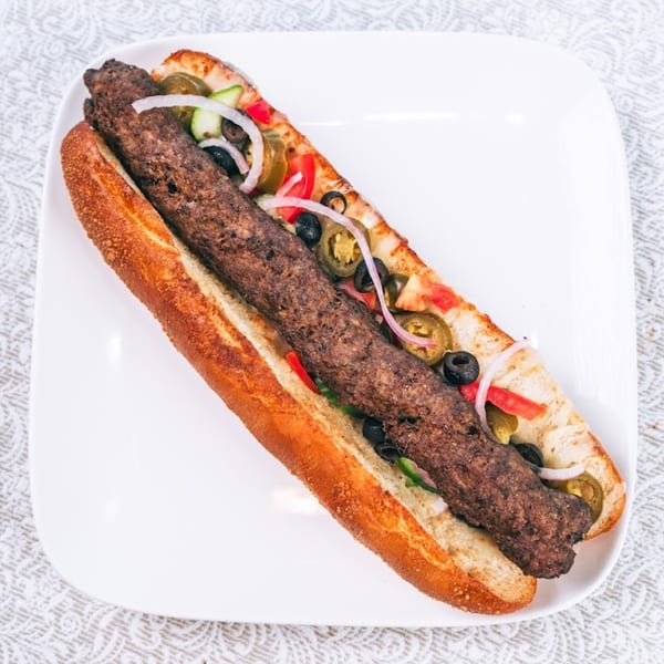 A long kebab sandwich with onions, olives, tomatoes, and jalapeños on a white plate.