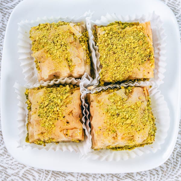 Four square pieces of baklava, topped with crushed pistachios, in white paper cups on a white plate.