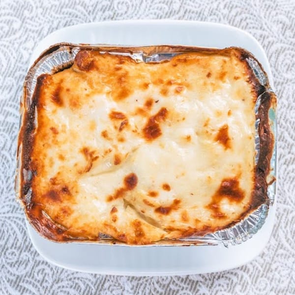 Baked lasagna in a foil container with a golden-brown top, served on a white plate.